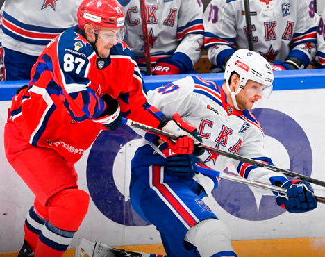 Хоккейный матч: игроки в форме красного и бело-синего цветов борются за шайбу у борта. На заднем плане скамейка запасных другой команды. Динамичная игровая ситуация