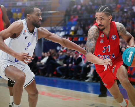 На изображении два баскетболиста в игровом моменте на баскетбольной площадке. Игрок в красной форме с мячом пытается обойти соперника в белой форме. На фоне видны зрители на трибунах